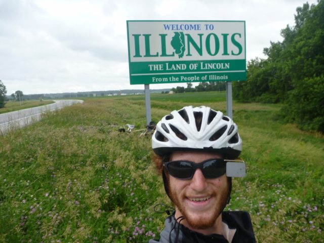 Michael Altfield stands in-front of a sign that reads "Welcome to Illonois" in USA