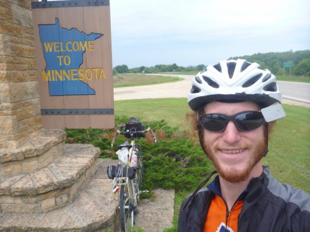 Michael Altfield stands in-front of a sign that reads "Welcome to Minnesota" in USA