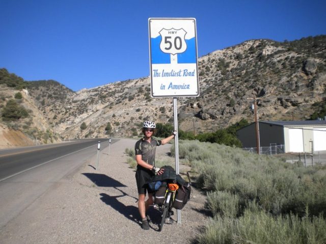 Michael Altfield stands in-front of a sign that reads "Hwy 50 The lonliest Road in America" in Nevada, USA.
