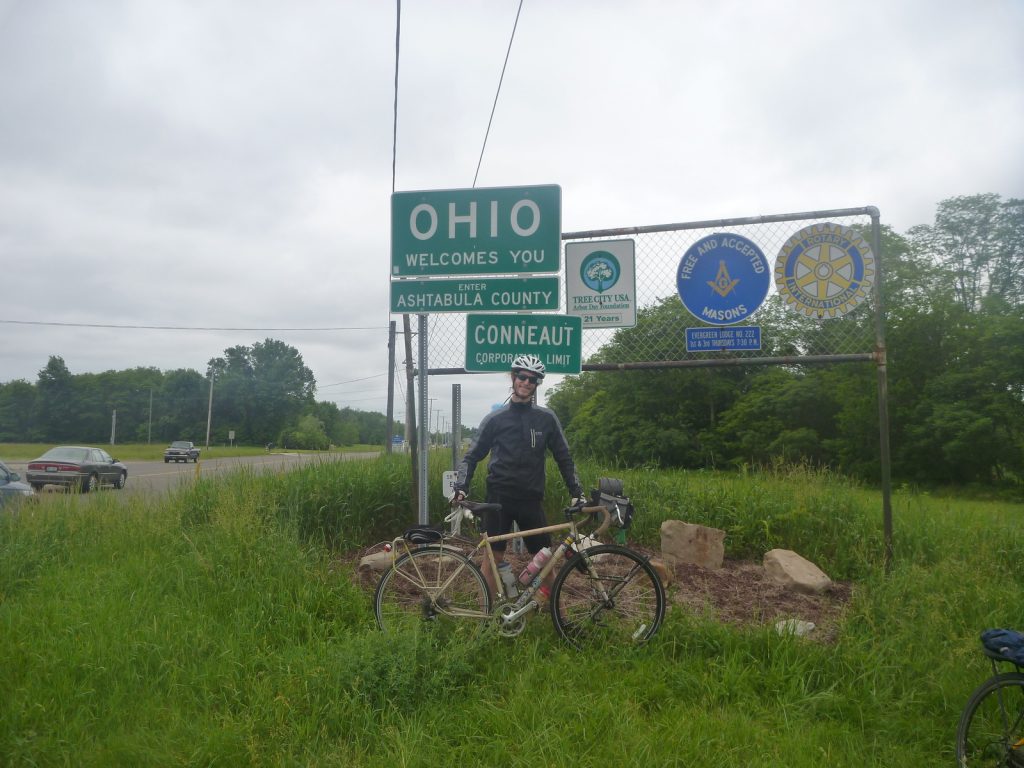 Michael Altfield stands with his bicycle in-front of a sign that says "Ohio Welcomes You" in USA